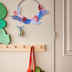 Studio roof pop out botanical wall decoration hanging on a beige wall above some wooden hangers and green leaves