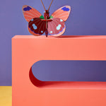 Studio roof eco-friendly delias butterfly slotting craft model balanced on a red sculpture block in front of a purple wall