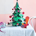Studio Roof Large cardboard Christmas Tree & cardboard Squirrel Decor in the front on a white draped table in front of a pink wall