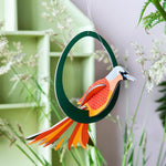 Close up of a Studio Roof renewable Paradise Bird Lady Obi decoration hanging from a plant stem in front of a green and purple background