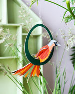 Close up of a Studio Roof renewable Paradise Bird Lady Obi decoration hanging from a plant stem in front of a green and purple background