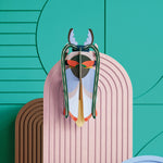 Studio roof slotting card cosmos beetle figure attached to a pink patterned panel in front of a green wall