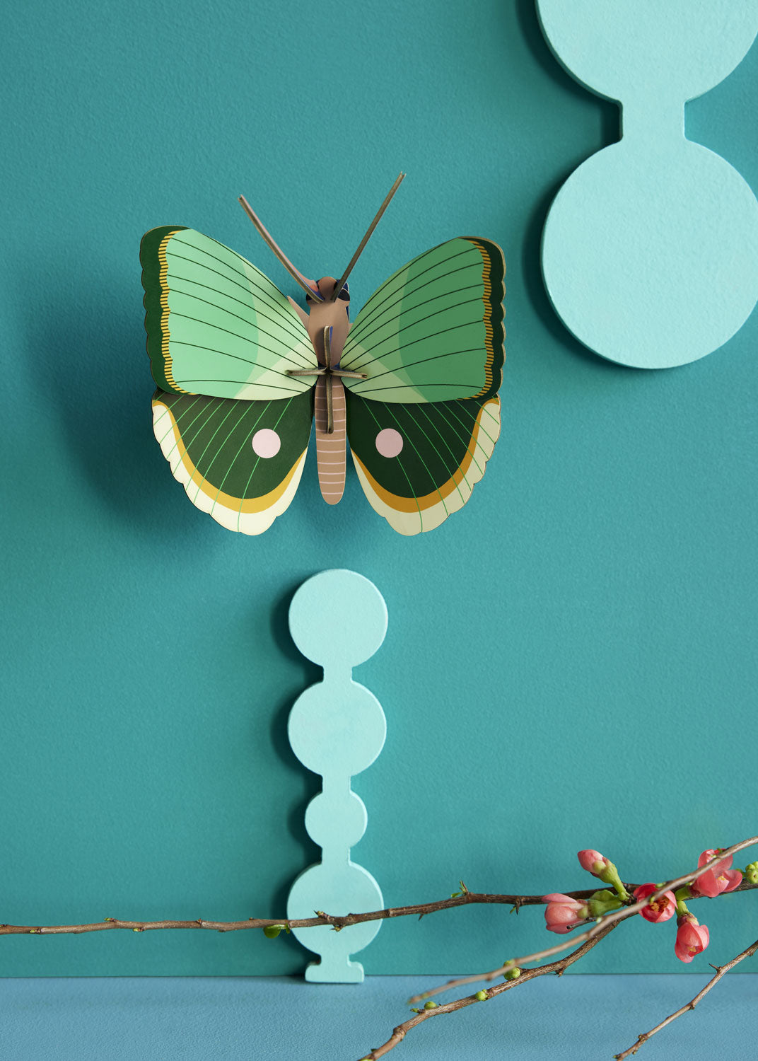 Studio Roof Fern Striped Butterfly decoration pictured on green blue painted wall with a branch with red flowers placed on a surface underneath