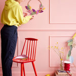 Woman stood next to a red wooden chair hanging a Studio roof eco-friendly blossom wreath decoration on a pink wall