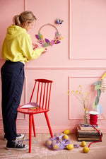 Woman stood next to a red wooden chair hanging a Studio roof eco-friendly blossom wreath decoration on a pink wall