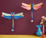 Studio Roof pink and blue dragonfly cardboard wall art kits shown built and displayed on a dark purple wall.