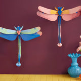 Studio Roof pink and blue dragonfly cardboard wall art kits shown built and displayed on a dark purple wall.