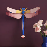 Studio Roof pink dragonfly cardboard wall art kit shown built and display on a dark purple wall.