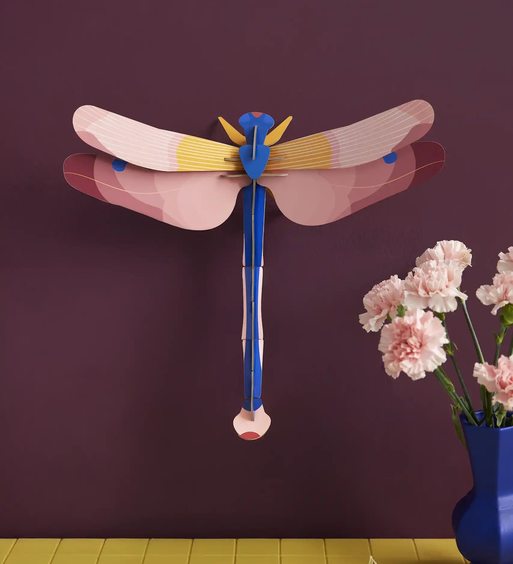 Studio Roof pink dragonfly cardboard wall art kit shown built and display on a dark purple wall.