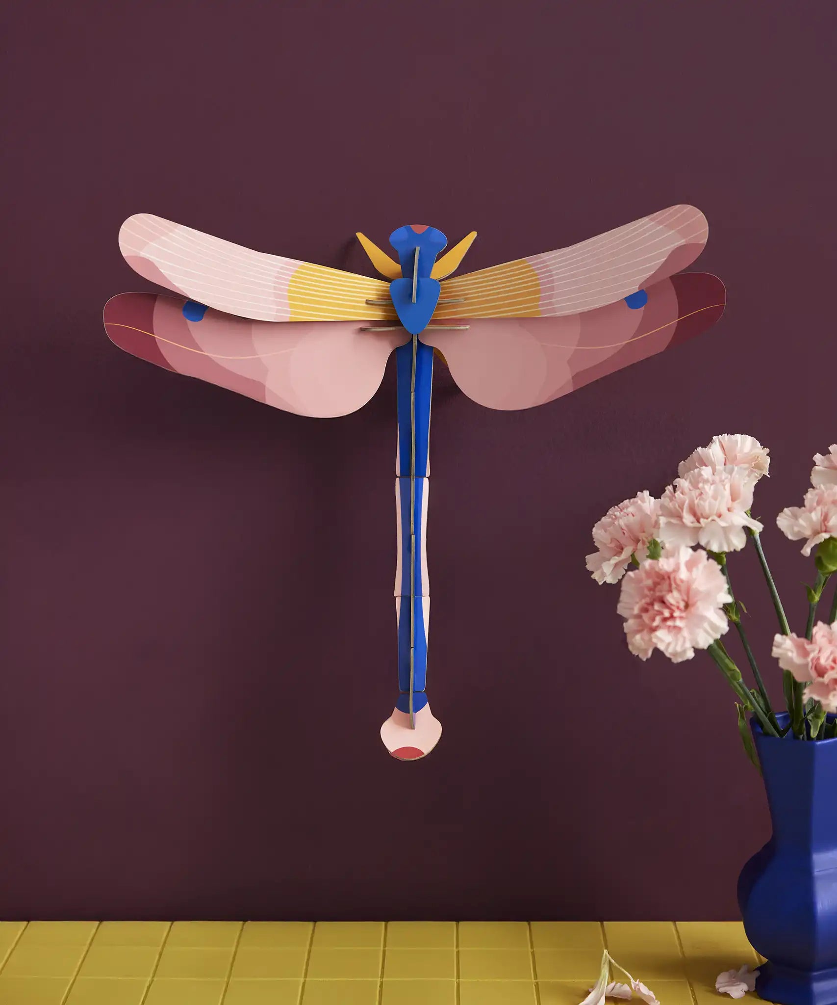 Studio Roof pink dragonfly cardboard wall art kit shown built and display on a dark purple wall.