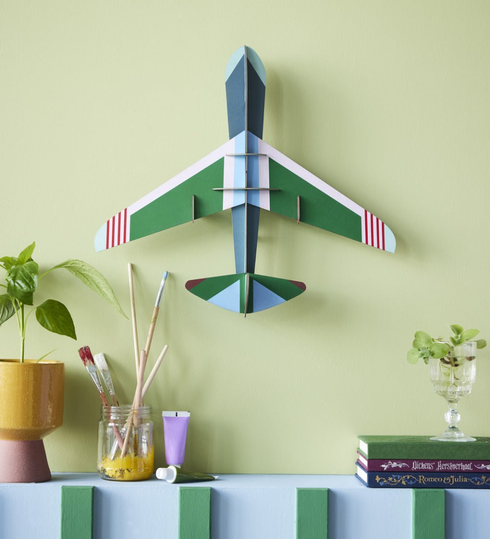 Studio Roof plastic-free card aeroplane on a green wall above a potted plant and some books