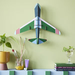 Studio Roof plastic-free card aeroplane on a green wall above a potted plant and some books