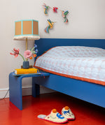 Studio ROOF Dance of Hummingbirds 3D cardboard wall art displayed above a bed, colourful hummingbird wall decorations flying around a flower in a bright bedroom setting.
