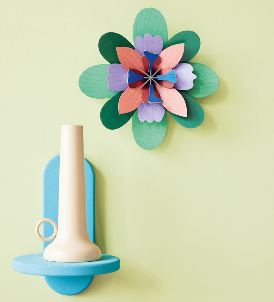 Studio Roof card craft flower wall hanging on a yellow wall next to a blue shelf and beige candle holder