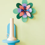 Studio Roof card craft flower wall hanging on a yellow wall next to a blue shelf and beige candle holder