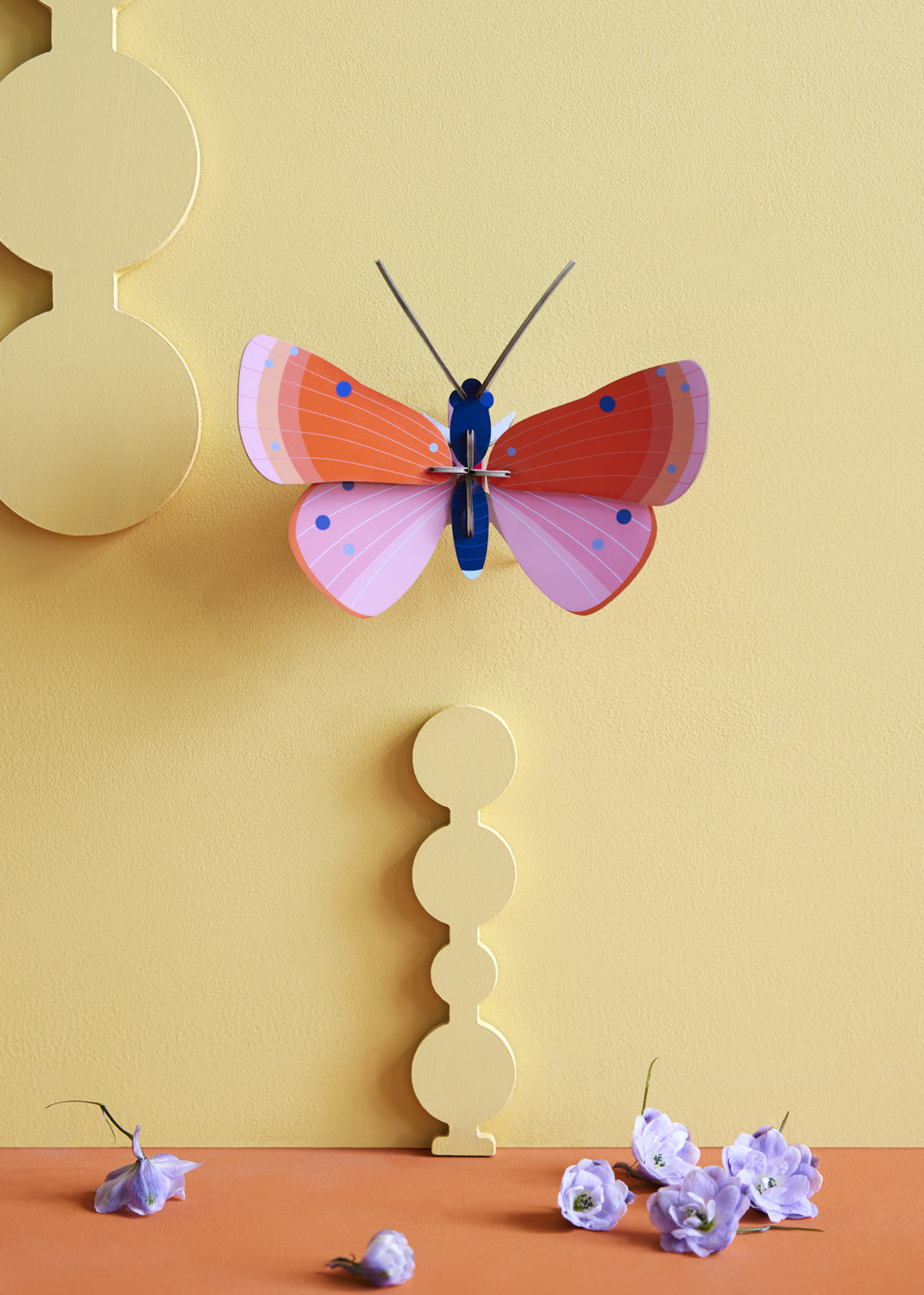 Studio Roof Speckled Copper Butterfly decoration on a pastel yellow coloured painted wall. Purple flowers can be seen scattered on a surface at the bottom