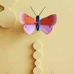 Studio Roof Speckled Copper Butterfly decoration on a pastel yellow coloured painted wall. Purple flowers can be seen scattered on a surface at the bottom
