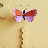 Studio Roof Speckled Copper Butterfly decoration on a pastel yellow coloured painted wall. Purple flowers can be seen scattered on a surface at the bottom