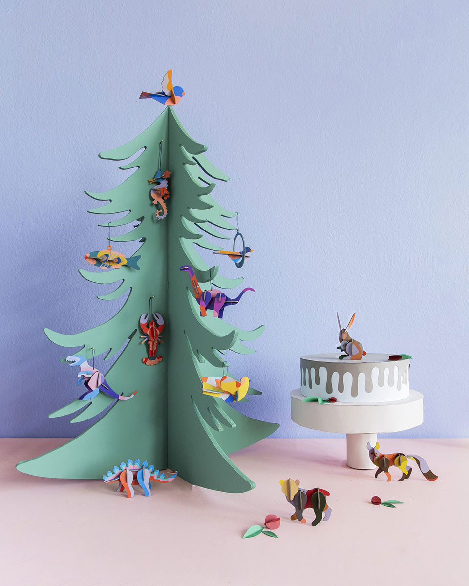 Studio Roof eco-friendly cardboard ornaments hanging from a wooden christmas tree in front of a blue wall