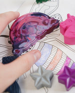 Close up of a hand reaching out to hold the Crayon Bug fantasy crayons on a colouring book
