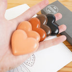 Close up of a hand holding the Crayon Bug skin tone heart crayons over a wooden background and colouring book