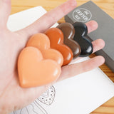 Close up of a hand holding the Crayon Bug skin tone heart crayons over a wooden background and colouring book