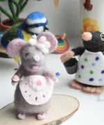 A felted mouse figure wearing a pink apron and holding a cake and lollypop in it's hands. The figure has been created using The Makerss Candy Mouse Needle Felt Craft Kit.