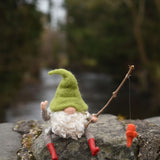 A felted gnome in a seated position with a fishing rod fashioned from a wooden stick created using The Makerss - Small Fishing Gnome Needle Felt Kit. 
