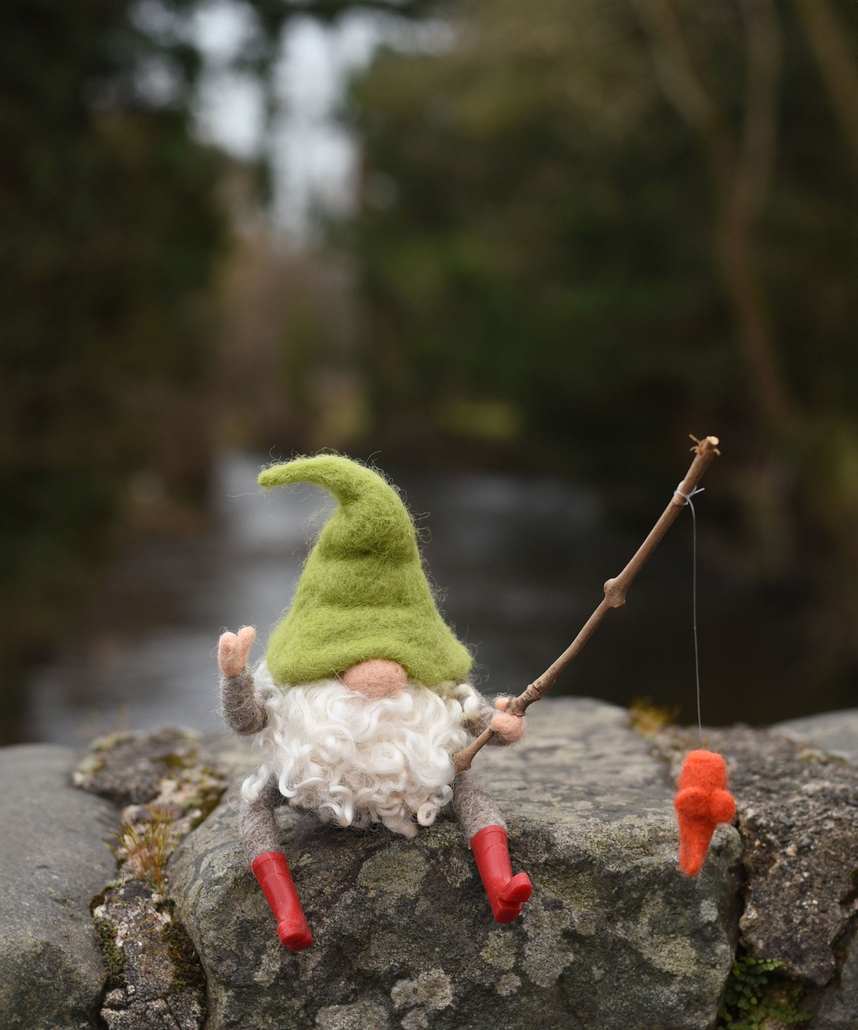 A felted gnome in a seated position with a fishing rod fashioned from a wooden stick created using The Makerss - Small Fishing Gnome Needle Felt Kit. 
