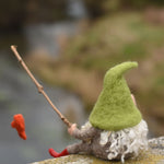 A felted gnome in a seated position with a fishing rod fashioned from a wooden stick created using The Makerss - Small Fishing Gnome. The gone is facing away from the camera revealing his long white wavy. hair
