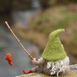 A felted gnome in a seated position with a fishing rod fashioned from a wooden stick created using The Makerss - Small Fishing Gnome. The gone is facing away from the camera revealing his long white wavy. hair