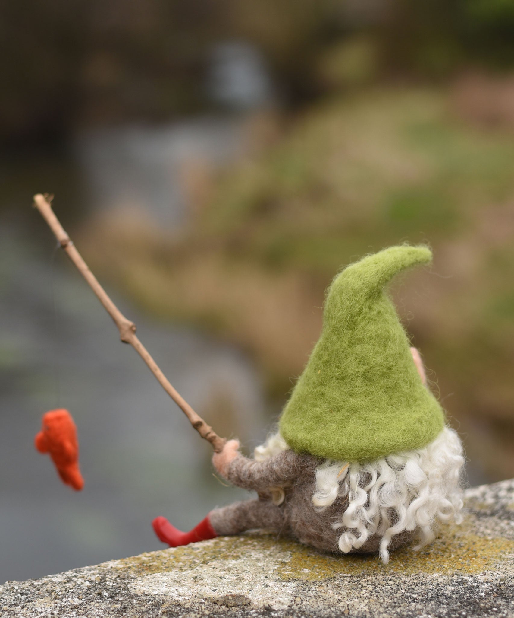 A felted gnome in a seated position with a fishing rod fashioned from a wooden stick created using The Makerss - Small Fishing Gnome. The gone is facing away from the camera revealing his long white wavy. hair