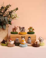 The Makerss - Amiguwoolli Tiny Mini Needle Felt Figures. 10 mini needle felt figures lined in two rows. Sat on top of a wooden box is the mini pig