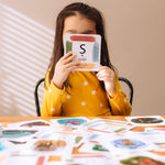 Child holding up a card with the letter S for snake on from the The Phive Enchanting Learning Phonics & Storytelling Cards - Set 1 with all the other cards laid out on the table in front of them