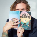 Author Dougie Poynter holding The Whale Watchers childrens book up in front of his face on a grey background