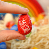 Close up of a hand holding a tonys chocolonely egg-stra special mini chocolate easter egg in front of a grimms wooden rainbow toy