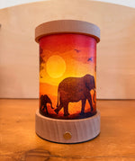 Toverlux Lantern shade by Vogel Geluk, transparency artworks of fauna  featuring elephants walking in a sunset. The shade has been displayed in a lit lantern. 