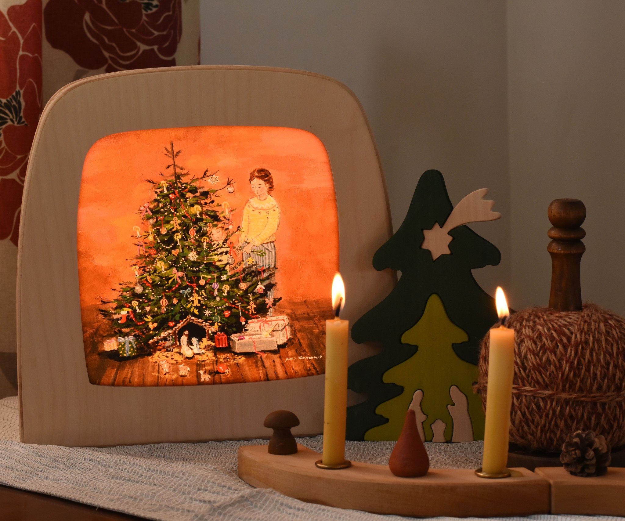 A Toverlux Magic Lamp with a Christmas tree Silhouette with artwork By Gwen van Knippenberg inside. The lamp is on and had a decorated Grimm's celebration ring placed in front of it.