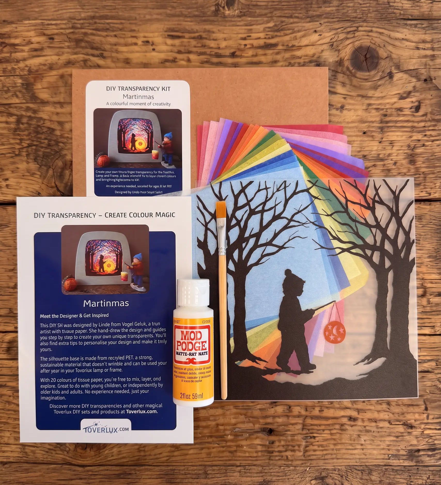 Toverlux Martinmas Silhouette DIY craft kit content laid out on a wooden surface. The kit includes silhouette panel with child carrying a lantern through a forest design, 20 sheets of coloured paper, paint brush, mod podge glue sealer and a step by step instruction guide booklet.