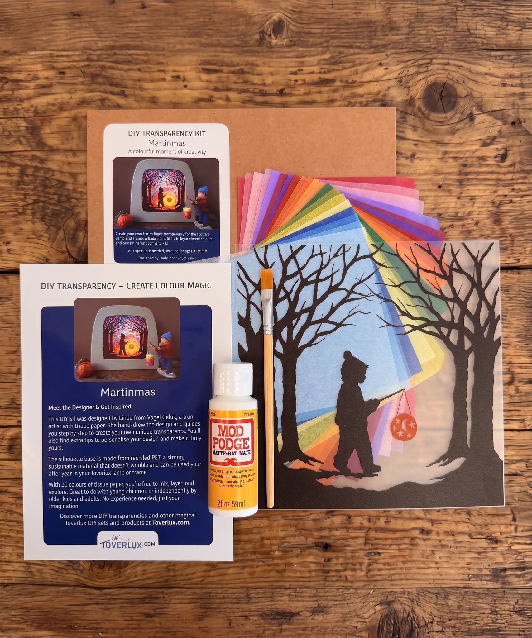 Toverlux Martinmas Silhouette DIY craft kit content laid out on a wooden surface. The kit includes silhouette panel with child carrying a lantern through a forest design, 20 sheets of coloured paper, paint brush, mod podge glue sealer and a step by step instruction guide booklet.