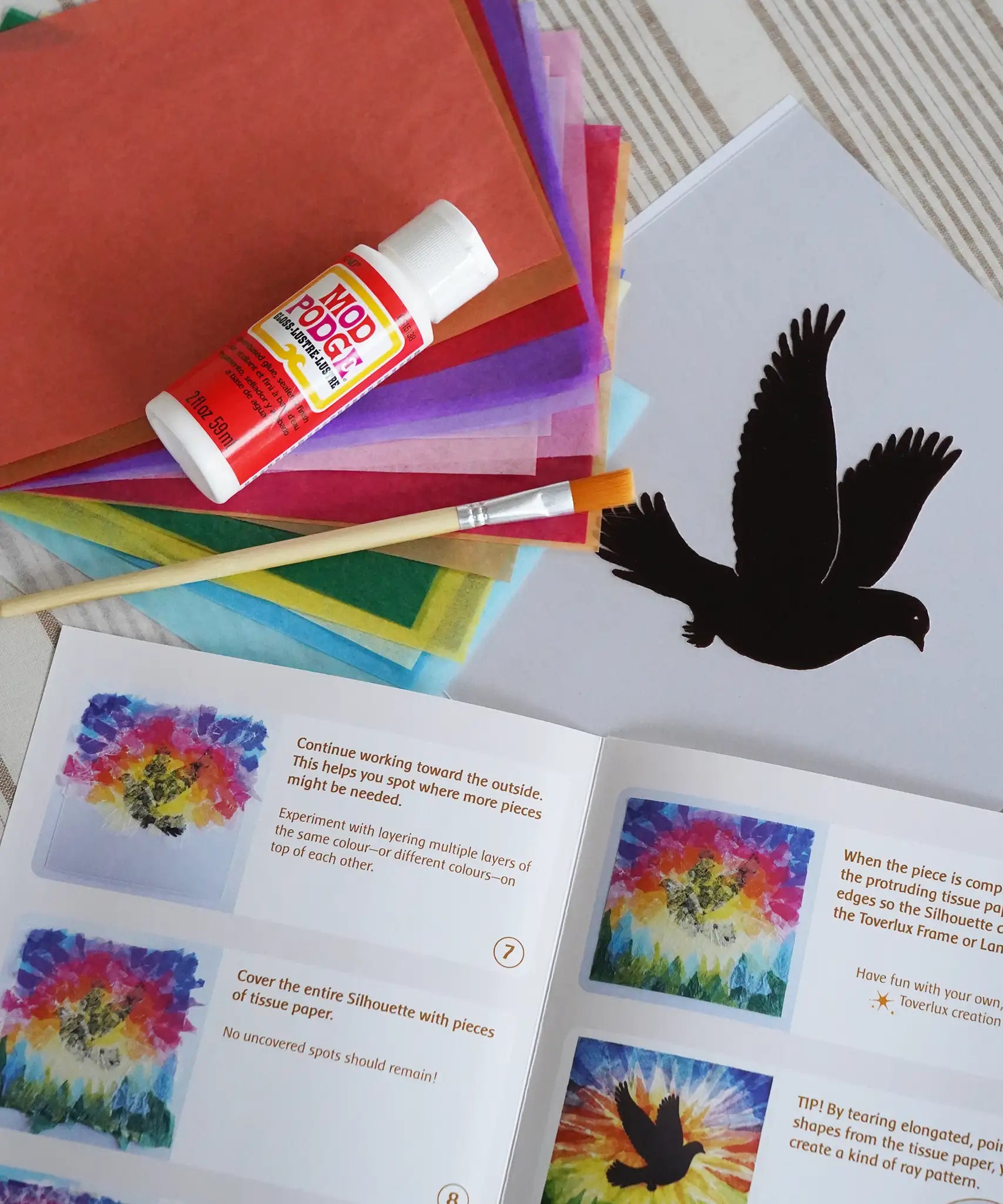 Contents of the Toverlux Spring dove DIY silhouette transparency craft kit featuring a Vogel Geluk design. The kit includes silhouette panel with dove design, 20 sheets of coloured paper, paint brush, mod podge glue sealer and a step by step instruction guide.