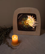 A warm cosy display with the Toverlux advent calendar inside a wooden lantern both available at Babipur. Lantern is standing on a table cloth next to a lit candle. 