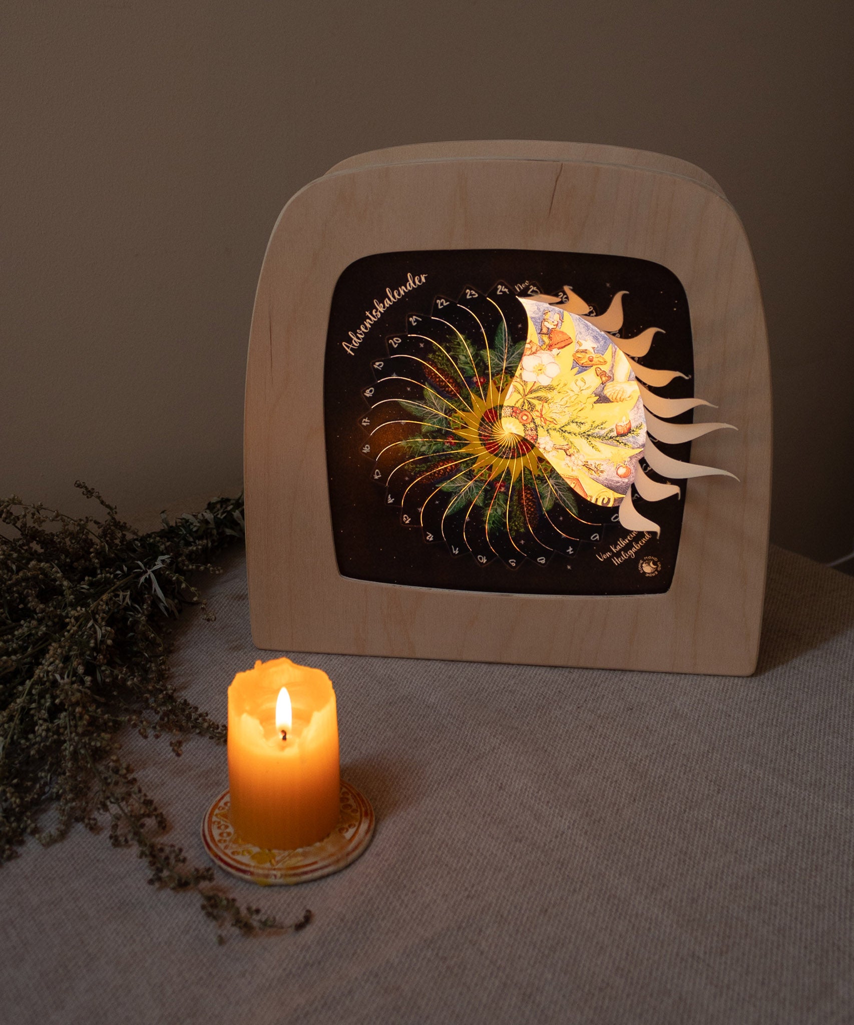 A warm cosy display with the Toverlux advent calendar inside a wooden lantern both available at Babipur. Lantern is standing on a table cloth next to a lit candle. 
