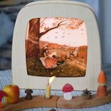 Autumn Lovers Picnic Toverlux Magic Lamp Silhouette placed in a Magic lamp on a table with a decorated Grimm's celebration ring in front of it. Artwork By Robin-Pieterse. A delicate painting style illustration of 2 people sitting underneath a tree. One of them is reading a book. A range of Toverlux silhouettes, lamp and accessories are available at Babipur.