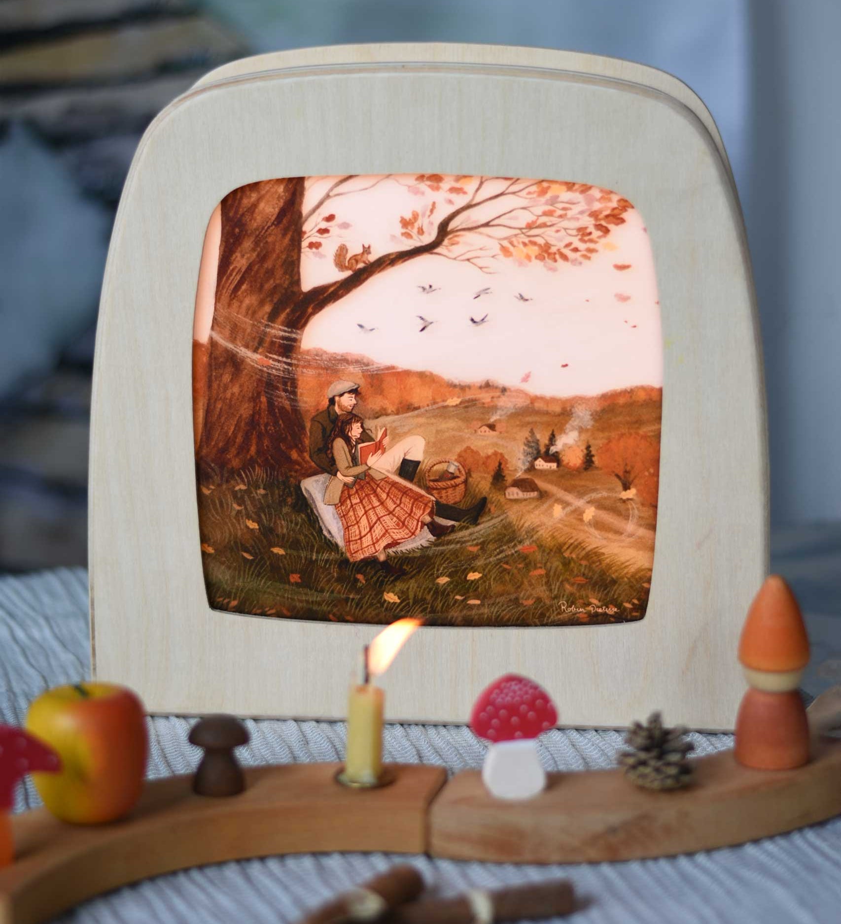 Autumn Lovers Picnic Toverlux Magic Lamp Silhouette placed in a Magic lamp on a table with a decorated Grimm's celebration ring in front of it. Artwork By Robin-Pieterse. A delicate painting style illustration of 2 people sitting underneath a tree. One of them is reading a book. A range of Toverlux silhouettes, lamp and accessories are available at Babipur.