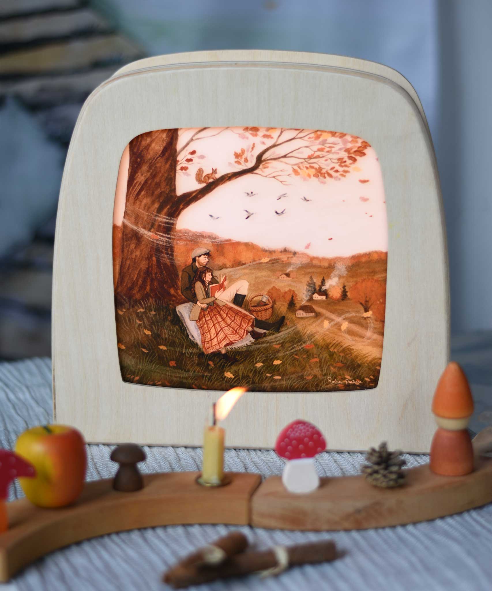 Autumn Lovers Picnic Toverlux Magic Lamp Silhouette placed in a Magic lamp on a table with a decorated Grimm's celebration ring in front of it. Artwork By Robin-Pieterse. A delicate painting style illustration of 2 people sitting underneath a tree. One of them is reading a book. A range of Toverlux silhouettes, lamp and accessories are available at Babipur.