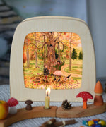 Toverlux Autumn Leaves Delight Toadstools Magic Lamp silhouette by artist Gwen van Knippenberg. The artwork has been placed in a magic lamp on a table with a decorated Grimm's celebration ring n front of it. A watercolour and pencil style illustration of children dancing under a tree whilst leaves are falling down around them.