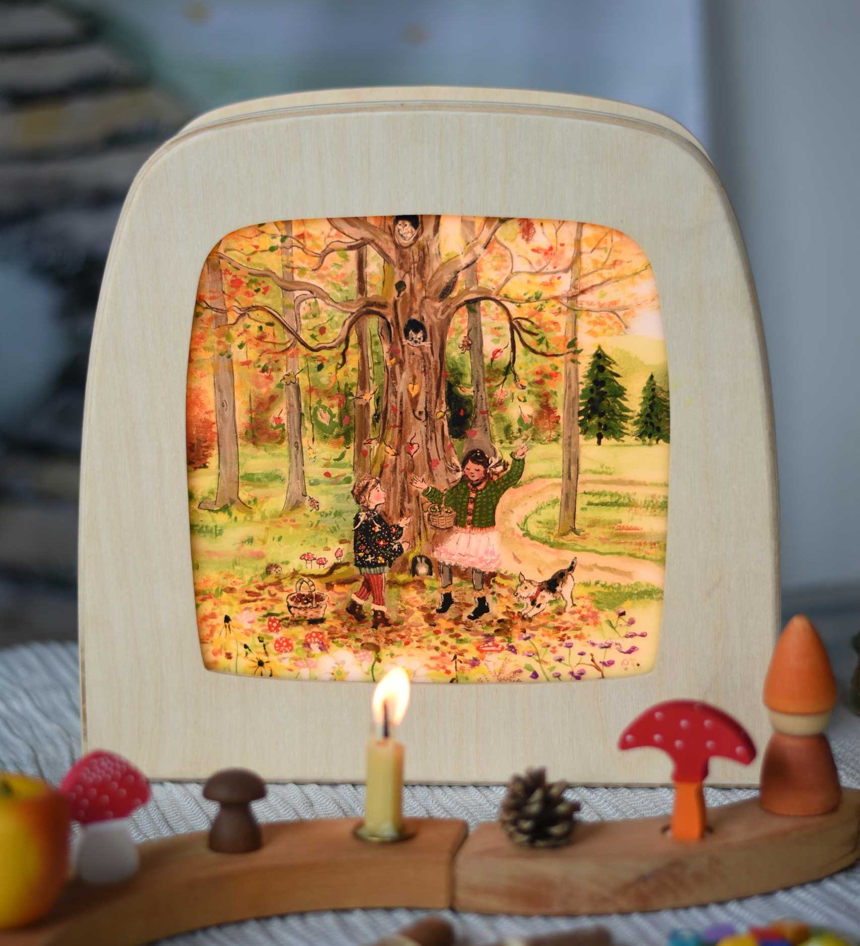 Toverlux Autumn Leaves Delight Toadstools Magic Lamp silhouette by artist Gwen van Knippenberg. The artwork has been placed in a magic lamp on a table with a decorated Grimm's celebration ring n front of it. A watercolour and pencil style illustration of children dancing under a tree whilst leaves are falling down around them.