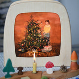 A Toverlux Christmas Tree Traditions Magic Lamp silhouette by artist Gwen van Knippenberg placed in a magic lamp on a table with a decorated Grimm's celebration ring in front of it. A watercolour and pencil style illustration of a woman and child standing by a decorated Christmas tree with candles on.