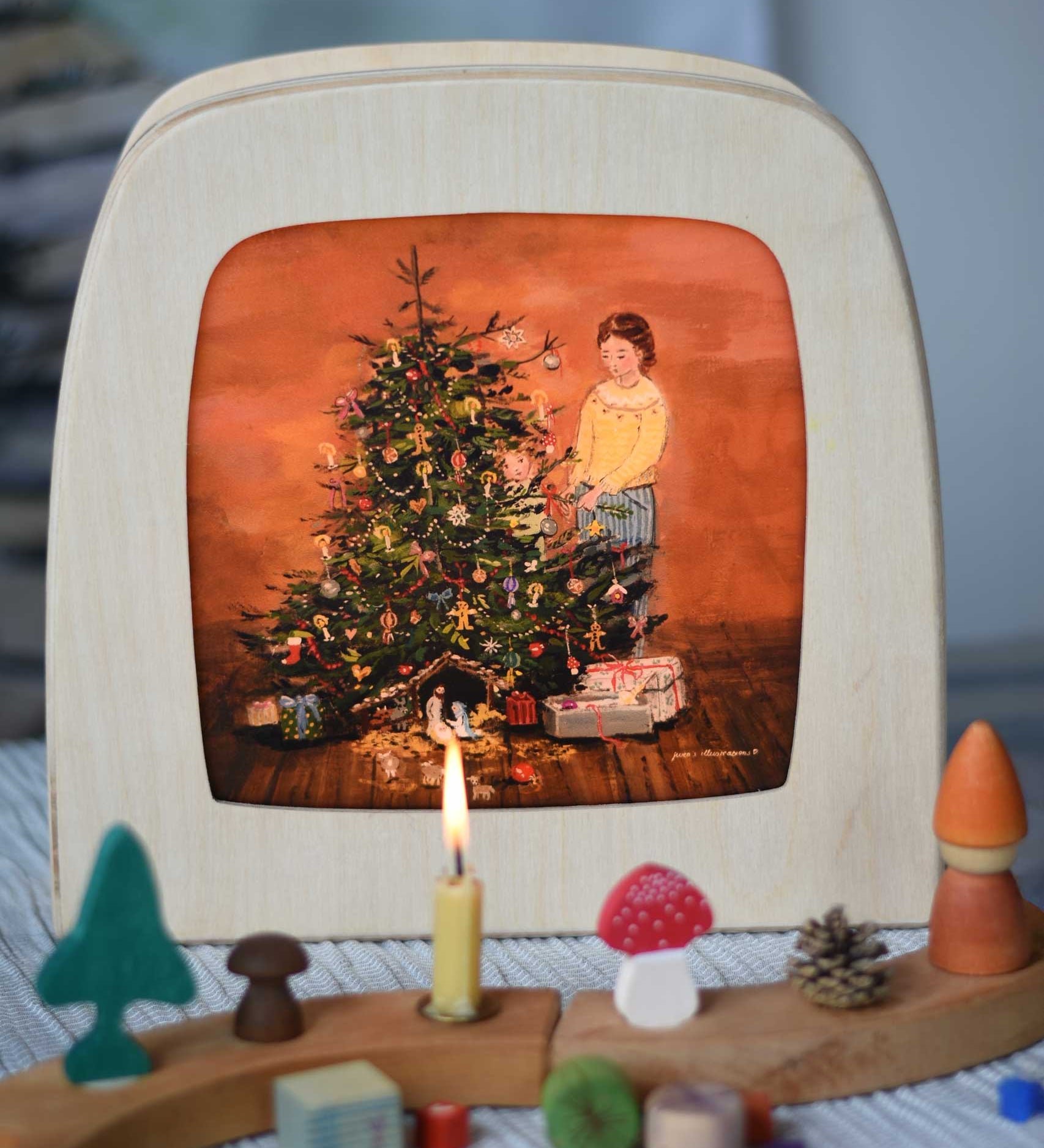 A Toverlux Christmas Tree Traditions Magic Lamp silhouette by artist Gwen van Knippenberg placed in a magic lamp on a table with a decorated Grimm's celebration ring in front of it. A watercolour and pencil style illustration of a woman and child standing by a decorated Christmas tree with candles on.